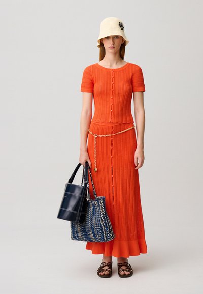 Mujer con top de punto naranja de manga corta con botones y falda larga a juego con cinturón de cadena dorada, sombrero beige, sandalias marrones, sosteniendo dos bolsos.