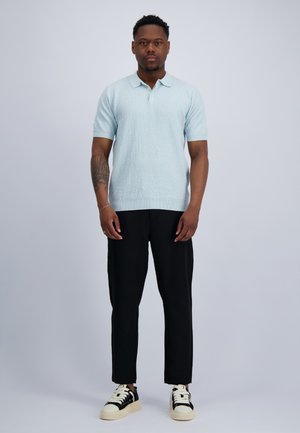 Man staat recht vooruit kijkend, draagt een lichtblauw poloshirt met korte mouwen, zwarte broek en zwart-witte sneakers tegen een effen grijze achtergrond.