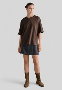 Brązowa, oversize koszulka z krótkim rękawem połączona z ciemną, kratkowaną, mini spódnicą i brązowymi, zamszowymi botkami za kostkę. Gładki materiał, luźny krój.