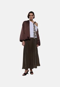 Giacca bomber in raso marrone, top bianco off-shoulder, sciarpa con stampa animale, gonna maxi plissettata oliva e décolleté aperti neri.