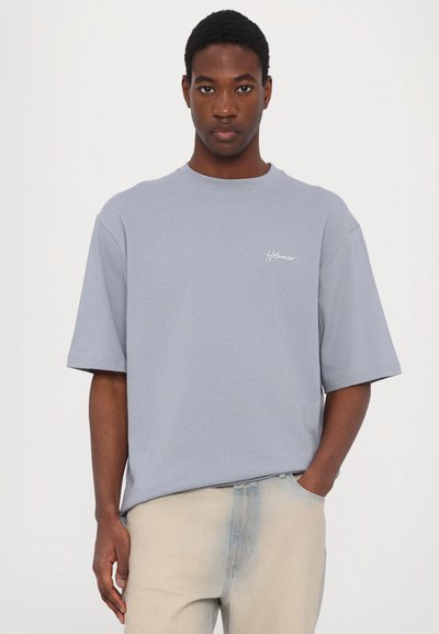 Light blue cotton t-shirt with short sleeves, round neckline, and a small white logo on the left chest. Worn with light-colored jeans.