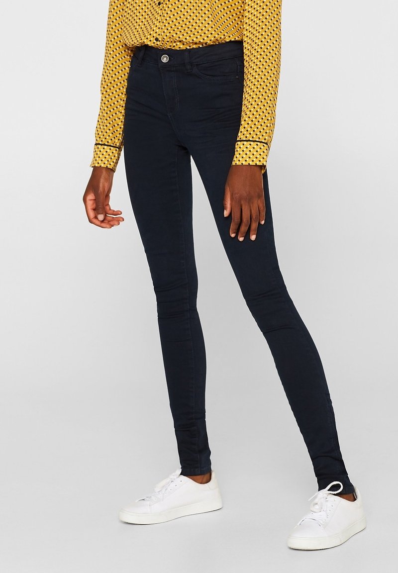 Jeans ajustados de color azul oscuro, hechos de denim elástico, con un cierre de botón estándar y bolsillos delanteros, acompañados de zapatillas deportivas blancas.