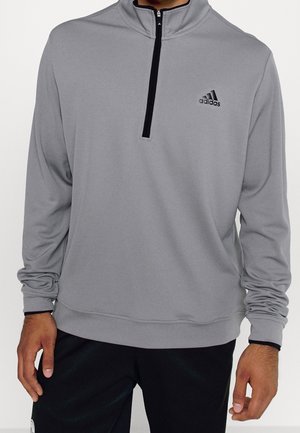 Mann trägt grauen Adidas Langarm-Pullover mit schwarzem Viertelreißverschluss und Logo auf der Brust, kombiniert mit schwarzen Hosen.