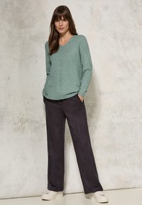 Mintgrön tröja med V-ringning, ribbade muddkanter och sidofickor, i kombination med löst sittande mörkgrå byxor och vita sneakers. Texturerad bakgrund.
