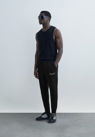 Άνδρας που στέκεται μπροστά σε λευκό τοίχο, φοράει μαύρο αμάνικο πουκάμισο, μαύρη φόρμα με λευκό λογότυπο, μαύρα και λευκά αθλητικά παπούτσια και μαύρα γυαλιά ηλίου.