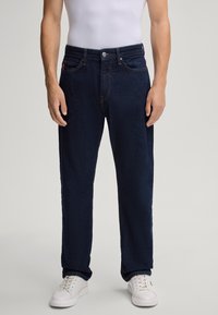 Dunkelblaue Jeans im geraden Schnitt, mit einem Fünf-Taschen-Design und einem Metallknopfverschluss. Getragen mit weißen Sneakers.