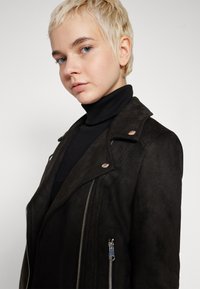 Zwarte suède motorjack met zilveren hardware, ritszakken en een gesneden kraag. Draagt over een zwarte turtleneck. Gladde textuur.