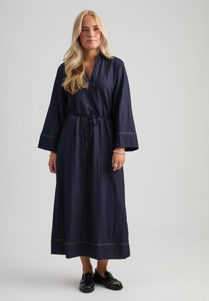Donna con lunghi capelli biondi che indossa un vestito midi a maniche lunghe blu scuro con cintura in vita e mocassini neri, in piedi davanti a uno sfondo semplice.