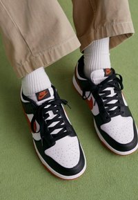 Les baskets Nike présentent un design noir et blanc avec des accents orange, une pointe perforée, des lacets noirs et une semelle texturée. Portées avec des chaussettes blanches.