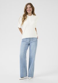 Witte polo met korte mouwen en een kraag, gecombineerd met lichtblauwe wijde jeans. Het model draagt puntige witte hakken. Eenvoudig ontwerp en pasvorm.
