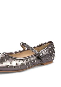 Zapato plano de plata metálica con puntera redonda, adornado con piedras de strass y una sola correa en el empeine asegurada con una hebilla decorativa.