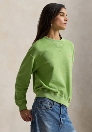 Felpa verde in cotone con scollo rotondo, maniche lunghe e piccolo logo rosso. Indossata con jeans di denim blu, mostrando una texture morbida.