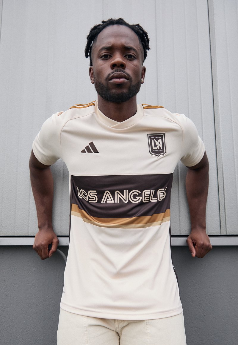 Maglietta da calcio bianca con strisce orizzontali marroni e dorate, testo "LOS ANGELES", logo Adidas e emblema della squadra. Texture liscia, vestibilità atletica.