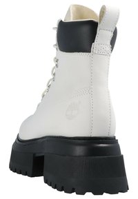 Botine albe din piele cu o talpă groasă din cauciuc negru, cu șireturi în față și un guler contrastant negru în partea de sus. Logo-ul Timberland este vizibil.