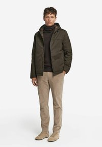 Ung mand iført en mørkebrun jakke, sort turtleneck, beige fløjlspant og beige ruskindsneakers, der står med hænderne i lommerne.