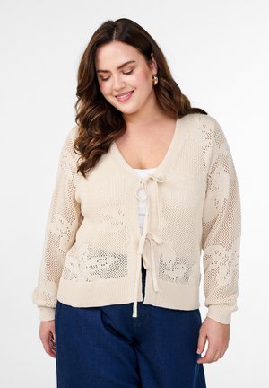 Cardigan lavorato a maglia in beige con motivo a trama aperta e chiusura a nodo sul davanti. Presenta maniche lunghe e polsini a coste, abbinato a un top bianco.