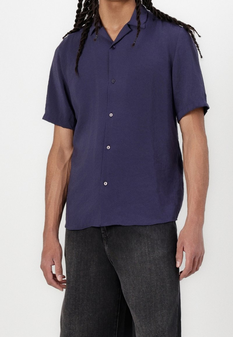 Homme portant une chemise bleu foncé à manches courtes boutonnée et un jean noir, debout devant un fond uni.