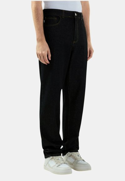 Persona che indossa jeans neri larghi e sneakers bianche, in piedi contro uno sfondo chiaro e uniforme.