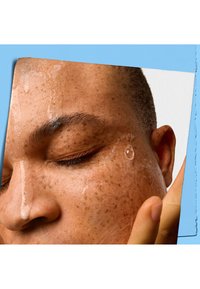 Nærbilde av et ansikt med våt hud, fremhevet av vanndråper. Synlige fregner, mørke øyenbryn og en nøytral bakgrunn med blå accenter.