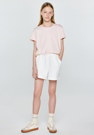 Młoda dziewczyna stoi z rękami w kieszeniach, ubrana w jasnoróżowy, paskowany t-shirt, białe szorty, kremowe skarpetki i beżowe trampki na szarym tle.