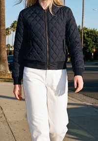 Personne marchant sur le trottoir portant une veste matelassée noire et un pantalon blanc avec des palmiers et une rue en arrière-plan.