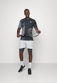 Camisa deportiva negra y gris con patrón a rayas a la izquierda; pantalones cortos grises con rayas negras; sosteniendo una raqueta de tenis; zapatos negros.