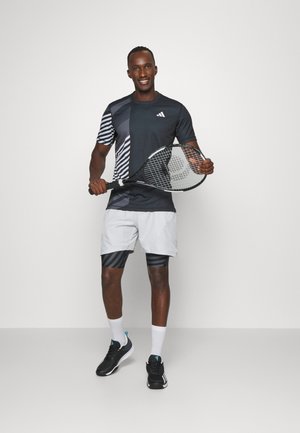 Camisa deportiva negra y gris con patrón a rayas a la izquierda; pantalones cortos grises con rayas negras; sosteniendo una raqueta de tenis; zapatos negros.