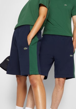 Zwei Personen tragen marineblaue Shorts mit grünen Seitenstreifen und grüne Hemden, die beide ein kleines Krokodillogo zeigen.