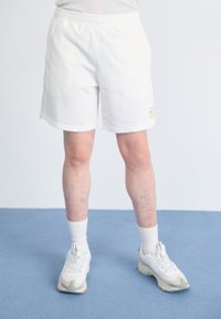 White athletic shorts with an elastic waistband and side pockets. Features a small beige logo. Worn with white sneakers and crew socks.
