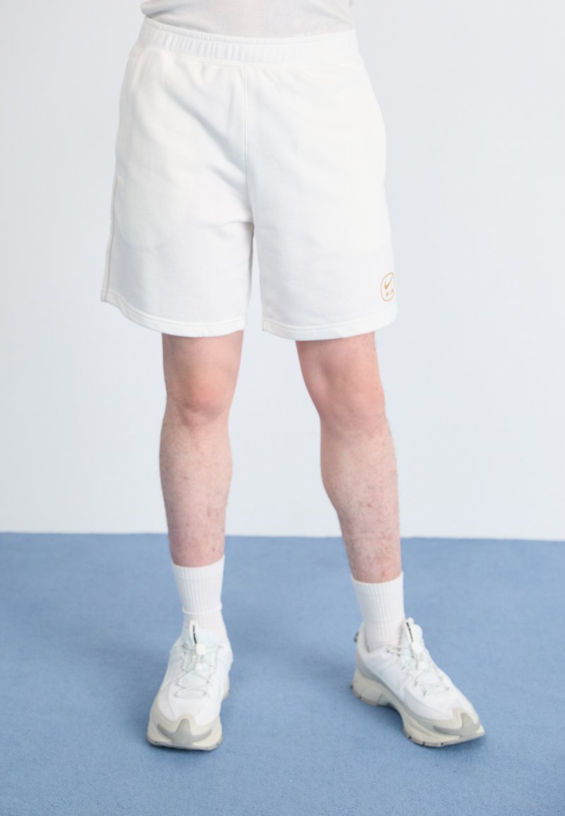 White athletic shorts with an elastic waistband and side pockets. Features a small beige logo. Worn with white sneakers and crew socks.