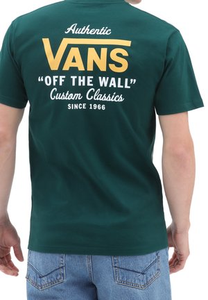 Persona che indossa una maglietta Vans verde scuro con testo giallo e bianco che recita "Authentic Vans Off The Wall Custom Classics Since 1966."