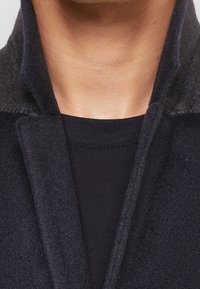 Cappotto di lana nero con una texture morbida, caratterizzato da un colletto a scialle e cuciture visibili. Indossato sopra una camicia nera, che mostra un look a strati.