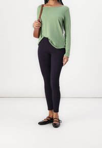 Grön långärmad topp med en lös passform, kombinerad med mörka leggings och svarta lägenheter med remmdetaljer. Enkel, avslappnad stil.