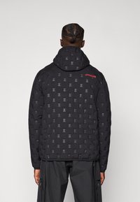 Black puffer jacket with a hood, featuring a geometric pattern, textured fabric, and a red logo accent on the upper back.