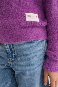 Lila bolyhos pulóver, amelyen a "JÓ DOLGOK JÖNNEK" feliratú címke található, világoskék farmernadrággal párosítva, amely csillogó díszítésekkel rendelkezik.