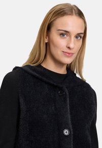 Schwarwest, flauschige Weste mit Kapuze und zwei Knöpfen. Der Stoff hat eine weiche Textur und ist über einem figurbetonten schwarzen Oberteil gestylt.