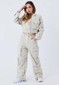 Chaqueta recortada beige con cremallera y múltiples bolsillos, combinada con pantalones cargo que tienen bolsillos laterales, ambos en una tela texturada. Zapatillas blancas.