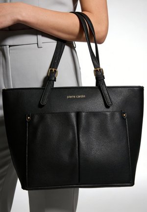 Zwarte leren tote bag met voorvakken en gouden gespen, gedragen door een persoon in lichtgrijze broek, zichtbaar "pierre cardin" logo.