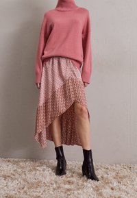 Rosa gestrickter Rollkragensweater, kombiniert mit einem gemusterten asymmetrischen Midikleid. Schwarze Knöchelstiefel runden das Outfit ab. Sanfte Textur, geschichteter Stil.