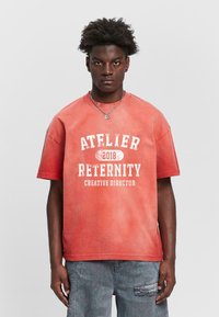 T-shirt en coton rouge avec un col rond, arborant le texte blanc "ATELIER RETERNITY 2018 DIRECTEUR CRÉATIF" dans une police vieillie. Coupe décontractée.
