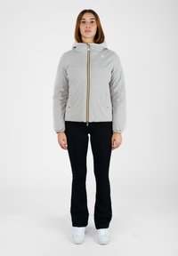 Giacca zip grigio chiaro con cappuccio, dotata di una cerniera frontale a righe multicolori, indossata sopra una maglietta nera e pantaloni, abbinata a sneakers bianche.