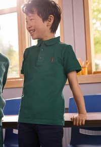 Grøn poloshirt med krave og tre knapper, med en rød hummel-logo på venstre bryst, båret sammen med mørke jeans.