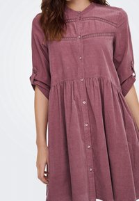Femme portant une robe boutonnée mauve avec des manches retroussées et un détail en dentelle au niveau de la poitrine et des épaules.