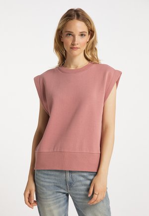Frau mit hellbraunen Haaren, die ein ärmelloses, staubig rosa Oberteil und hellblaue Jeans trägt, steht vor einem schlichten weißen Hintergrund.