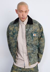 Chaqueta de lona verde con patrón de camuflaje, cuello negro, cierre de cremallera y parche de logo; llevada sobre una camisa de botones de color beige claro.