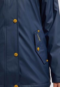 Námořnicky modrý nepromokavý plášť se žlutými knoflíky, obsahující boční kapse a hladkou texturou. Strukturovaný střih s knoflíkovým zapínáním vpředu.