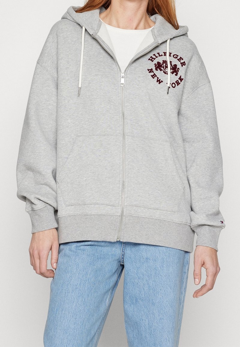 Personne portant un sweat à capuche zippé gris clair avec poches avant et emblème bordeaux "Hilfiger New York", associé à un jean bleu clair.