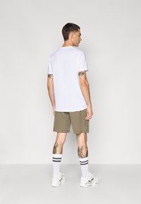 T-shirt branca de mangas curtas, calções cáqui, meias brancas com riscas pretas e ténis brancos; exibido visto de costas.