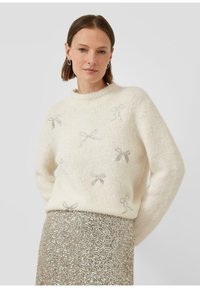Weißer gestrickter Pullover mit strukturiertem Finish und silbernen Schleifenmustern, kombiniert mit einem goldenem Rock aus Pailletten. Rundhalsausschnitt und lockere Passform.