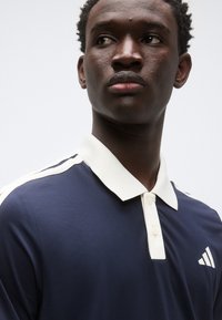 Polo bleu marine avec un col crème, doté de trois boutons et de bandes blanches sur les épaules. Affichant le logo Adidas sur le côté gauche.